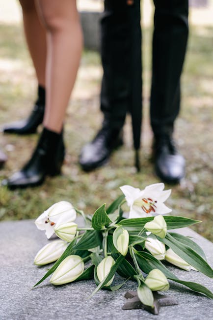 A bouquet of fresh white lilies, arranged with green leaves, rests on a stone surface outdoors with a blurred background showing the lower legs and feet of two people dressed formally in dark suits an